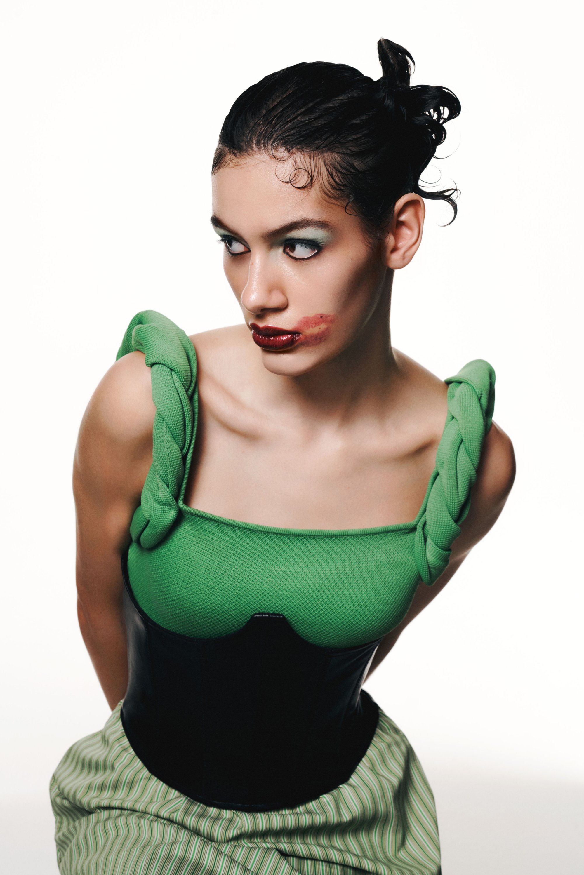 Una modelo con cabello oscuro recogido en dos moños lleva una blusa verde con tirantes trenzados y un corsé negro, con un maquillaje llamativo y una expresión dramática.