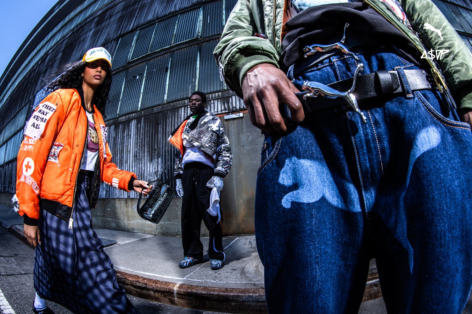 A woman in an orange bomber jacket walks with a shiny bag, while a man in a patterned jacket poses nearby against a textured wall.
