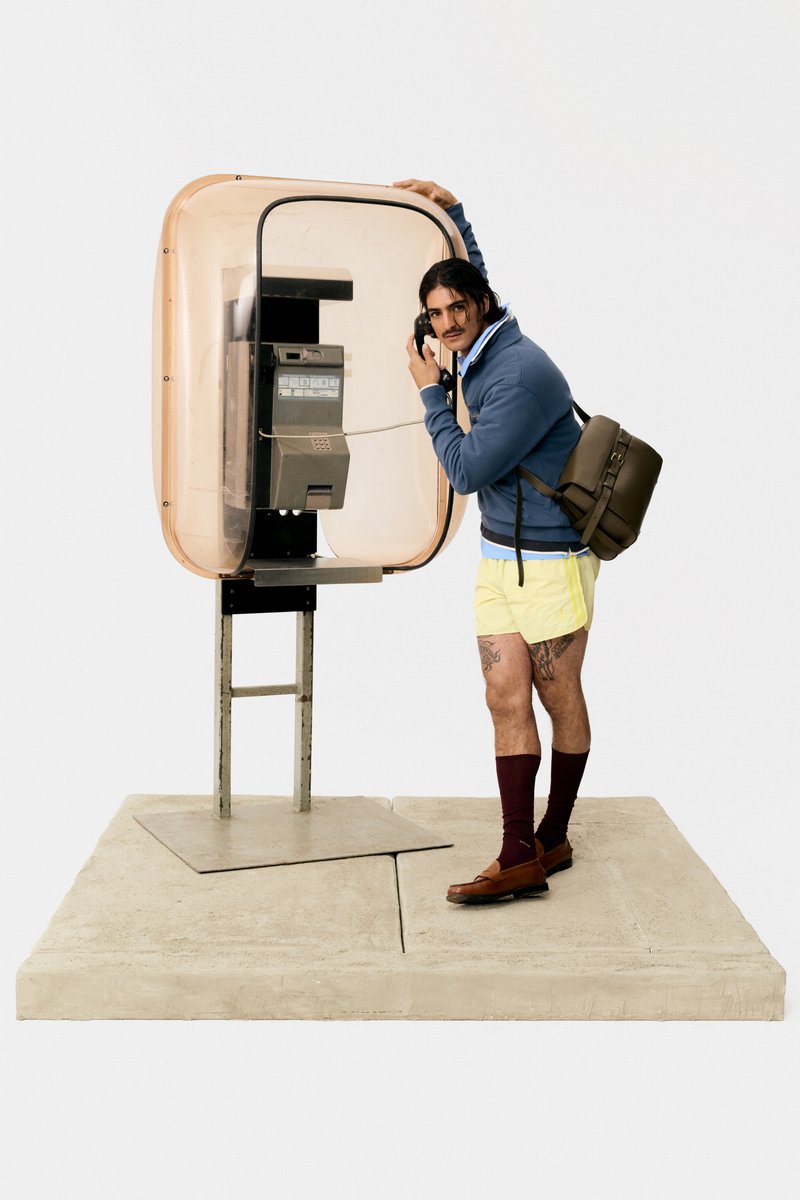 A man in yellow shorts and a blue hoodie talks on a payphone, leaning playfully against its translucent enclosure, with a backpack slung over one shoulder.