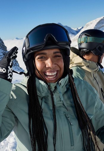 Γυναίκα φοράει ένα μπουφάν σκι σε μέντα πράσινο χρώμα, μαύρα γάντια με λευκές λεπτομέρειες και ένα μαύρο κράνος σκι με γυαλιά ηλίου σε μπλε απόχρωση σε χιονισμένα βουνά.