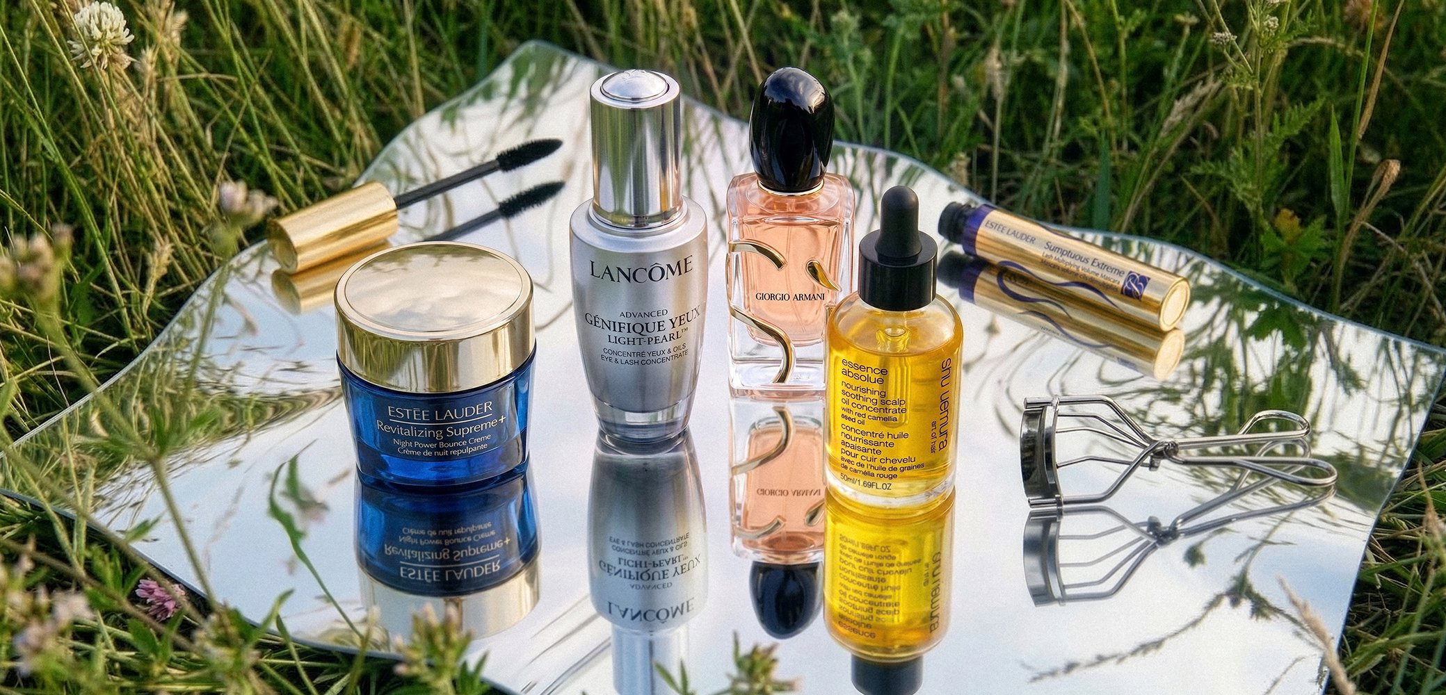 Skincare and makeup products including Estee Lauder cream, Lancôme serum, Giorgio Armani perfume, facial oil, mascara, and an eyelash curler on reflective surface outdoors.