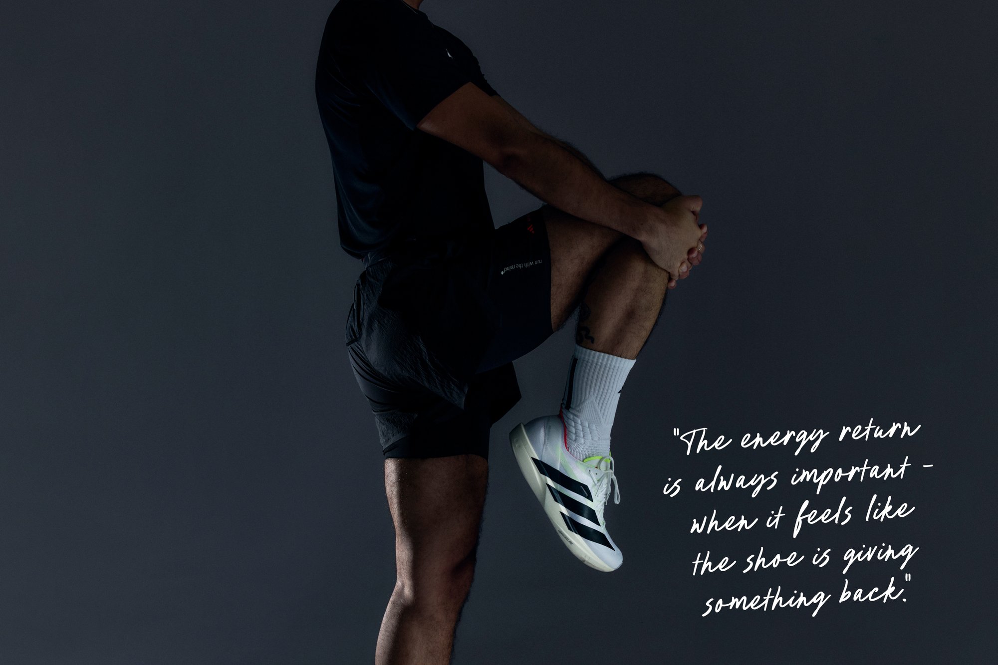 Schwarzes sportliches Kurzarmshirt und Shorts, weiße und schwarze Sneakers mit neonfarbenen Akzenten, sitzende Pose vor einem dunklen Hintergrund.