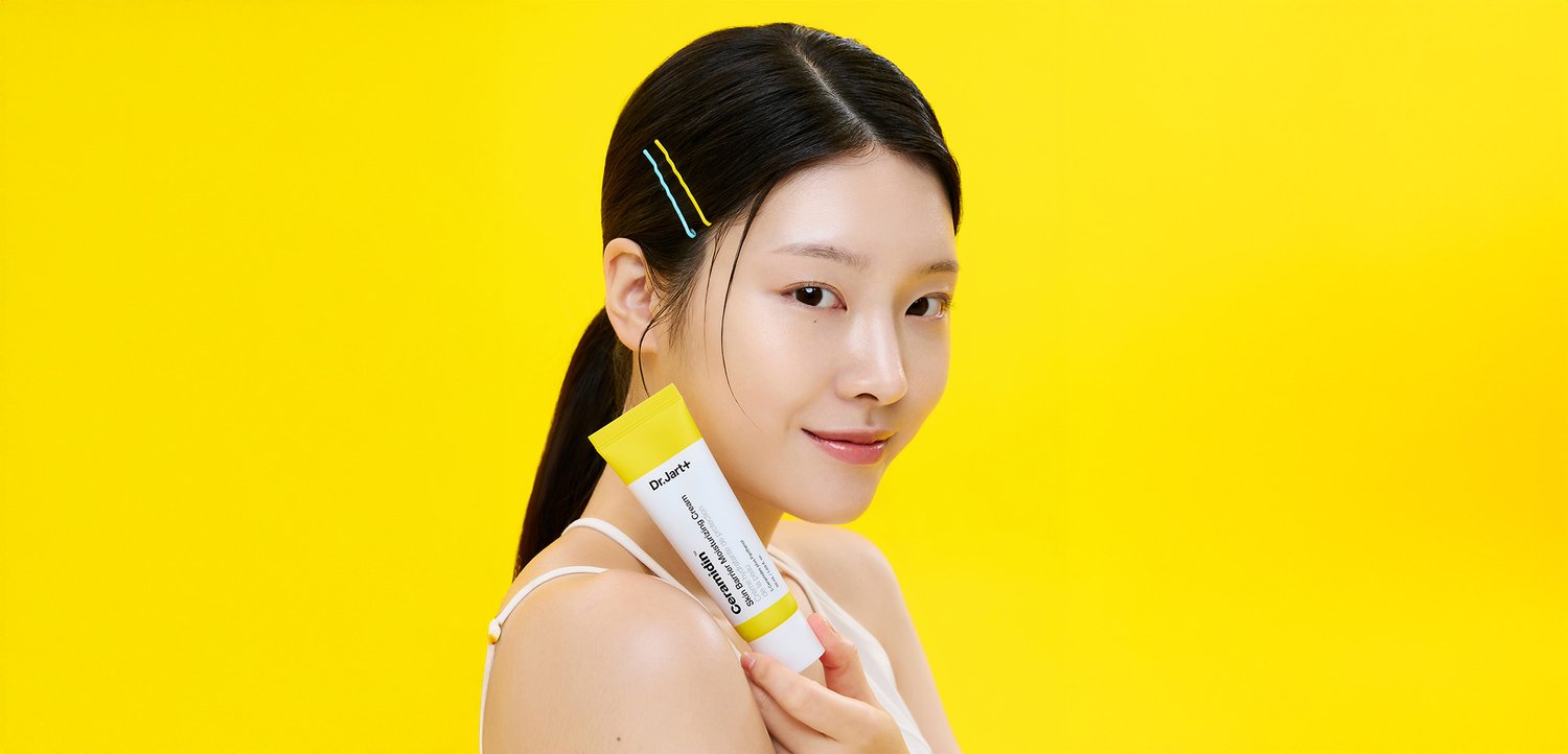 Dr. Jart+ Feuchtigkeitscreme in einer weißen und gelben Tube, gehalten von einer Person mit dunklen Haaren, vor einem einfarbigen gelben Hintergrund.