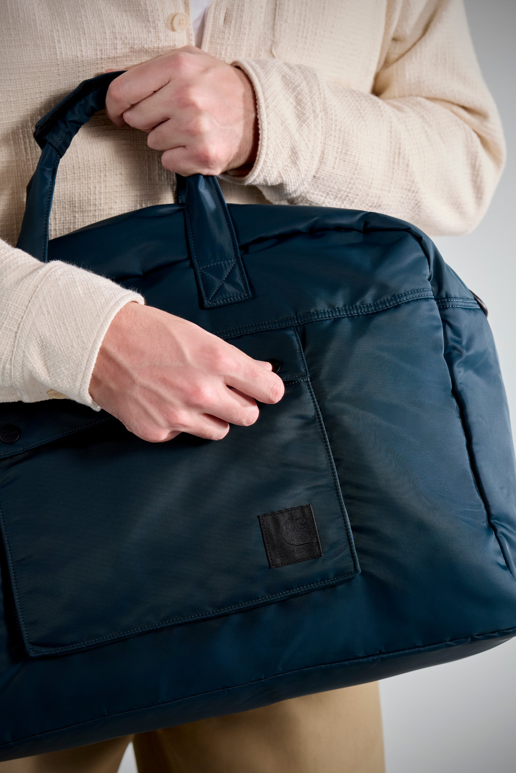 A hand grips the handle of a dark blue bag with a small pocket, worn by a person in a cream sweater against a neutral background.