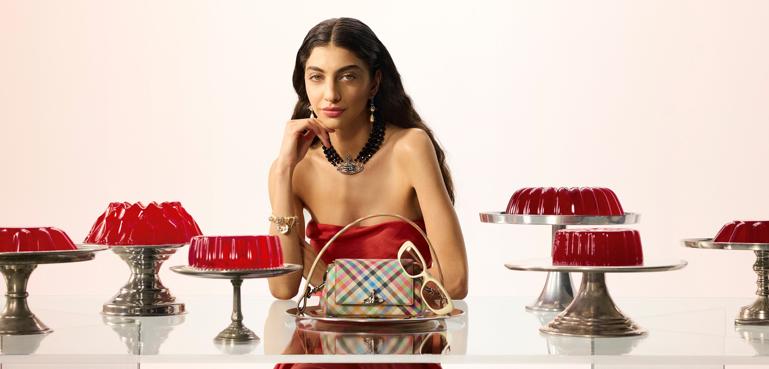 Mujer con vestido rojo sin tirantes sentada en una mesa con moldes de gelatina roja sobre soportes de plata, sosteniendo un bolso de cuadros y unas gafas de sol.