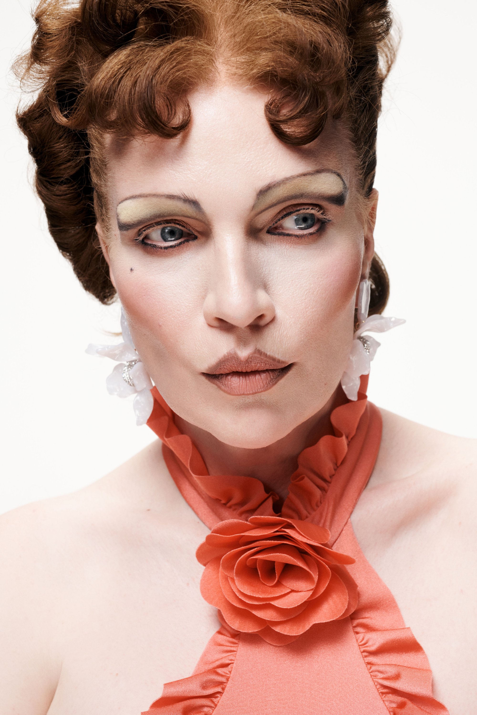 A woman with styled, curly hair and dramatic makeup wears an orange ruffled top adorned with a fabric rose. Elegantly posed against a white background.