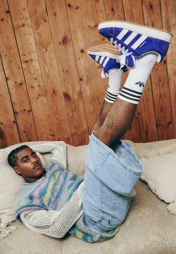 Eine Person liegt entspannt auf einem Sofa mit hochgelegten Beinen, trägt einen bunten Pullover und zerrissene Jeans und zeigt lebhafte blaue Sneakers und gestreifte Socken.