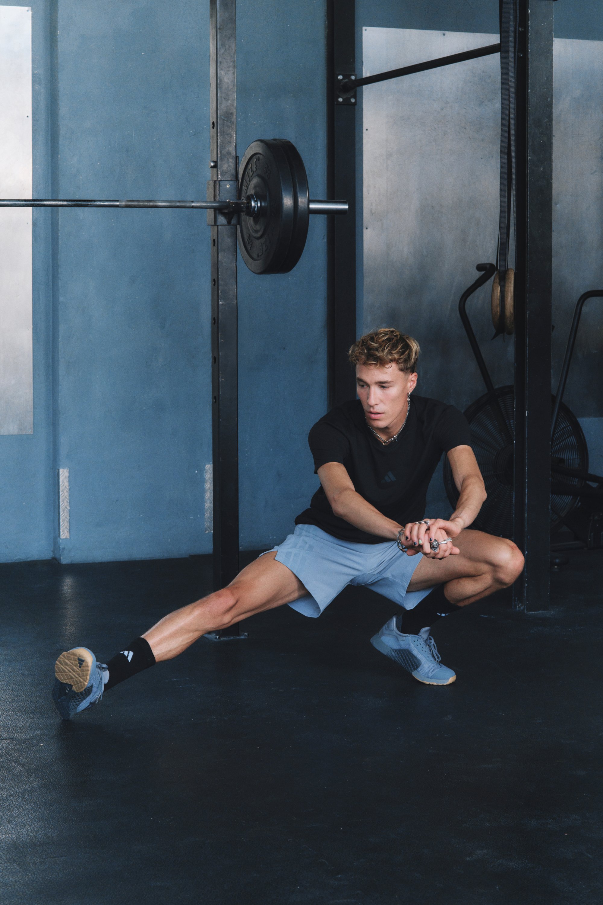 Atleet in zwart shirt en lichtgrijze short voert een atletische stretch uit. De omgeving bevat metalen fitnessapparatuur en een blauwe muur.