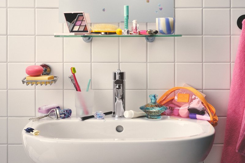Badeværelsesvask med vandhane, tandbørste i matteret holder, sæbestykker, makeup-palet, hårbørste, parfume flaske, og pink håndklæde på flisebelagt væg baggrund.