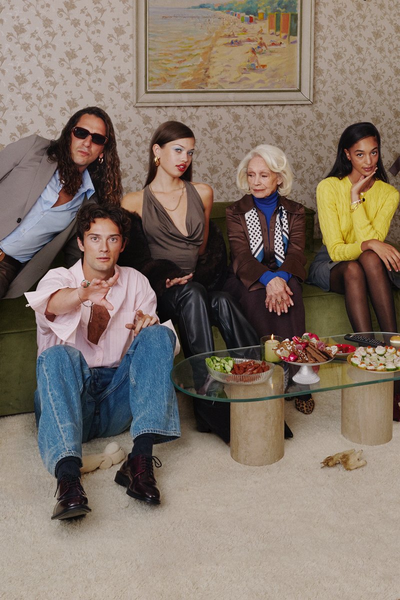 Grupo de cinco personas sentadas en un sofá verde con una alfombra blanca y peluda, mesa de café de vidrio con aperitivos, papel pintado con patrón y pintura de playa.