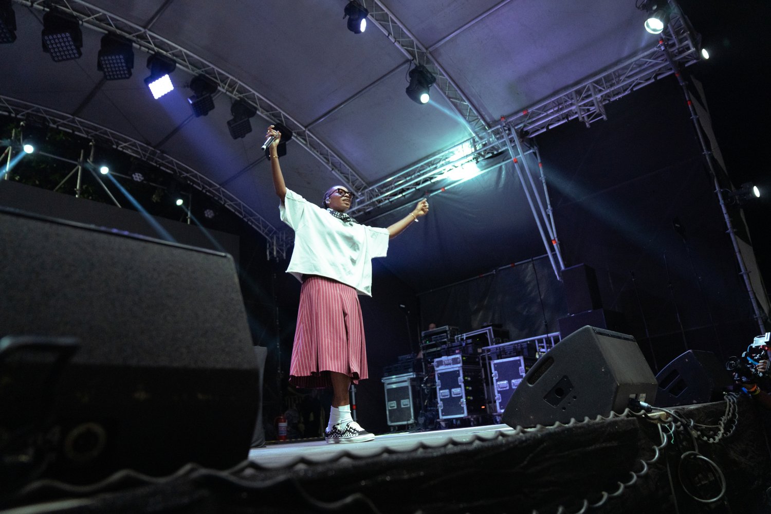 A performer in a loose white top and pink striped skirt holds a microphone, arms raised, on stage with colorful lights and audience behind.