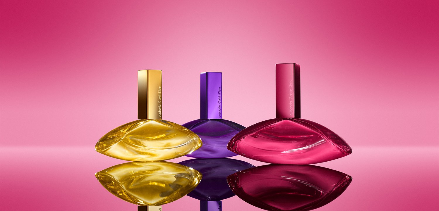 Tre buede parfume flasker formet som blade i gul, lilla og pink med matchende låg, reflekteret på en blank pink overflade.