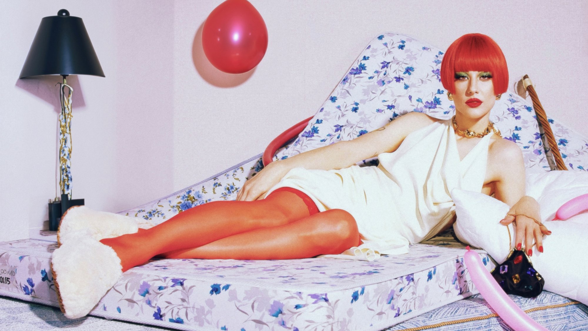 A person with a red bob haircut lounges on a floral mattress, wearing a white dress and knee-high red stockings, surrounded by decorative pillows and balloons.