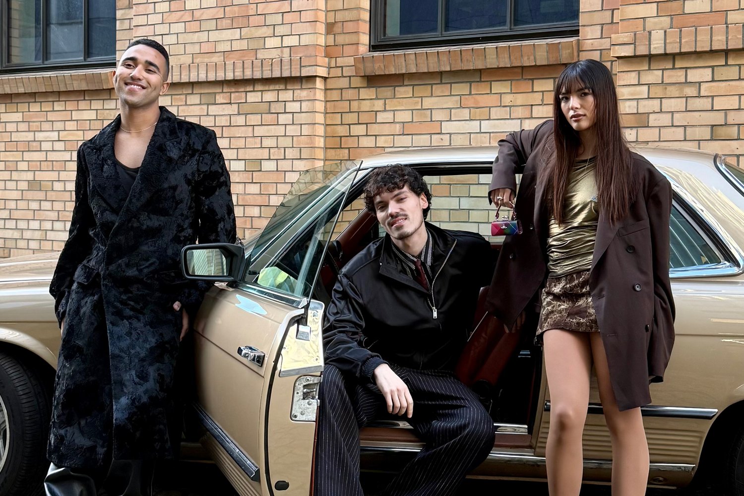 Tre persone alla moda posano accanto a un'auto d'epoca: un uomo sorridente in un cappotto nero, un uomo seduto in giacca e una donna in mini gonna che tiene una borsa.