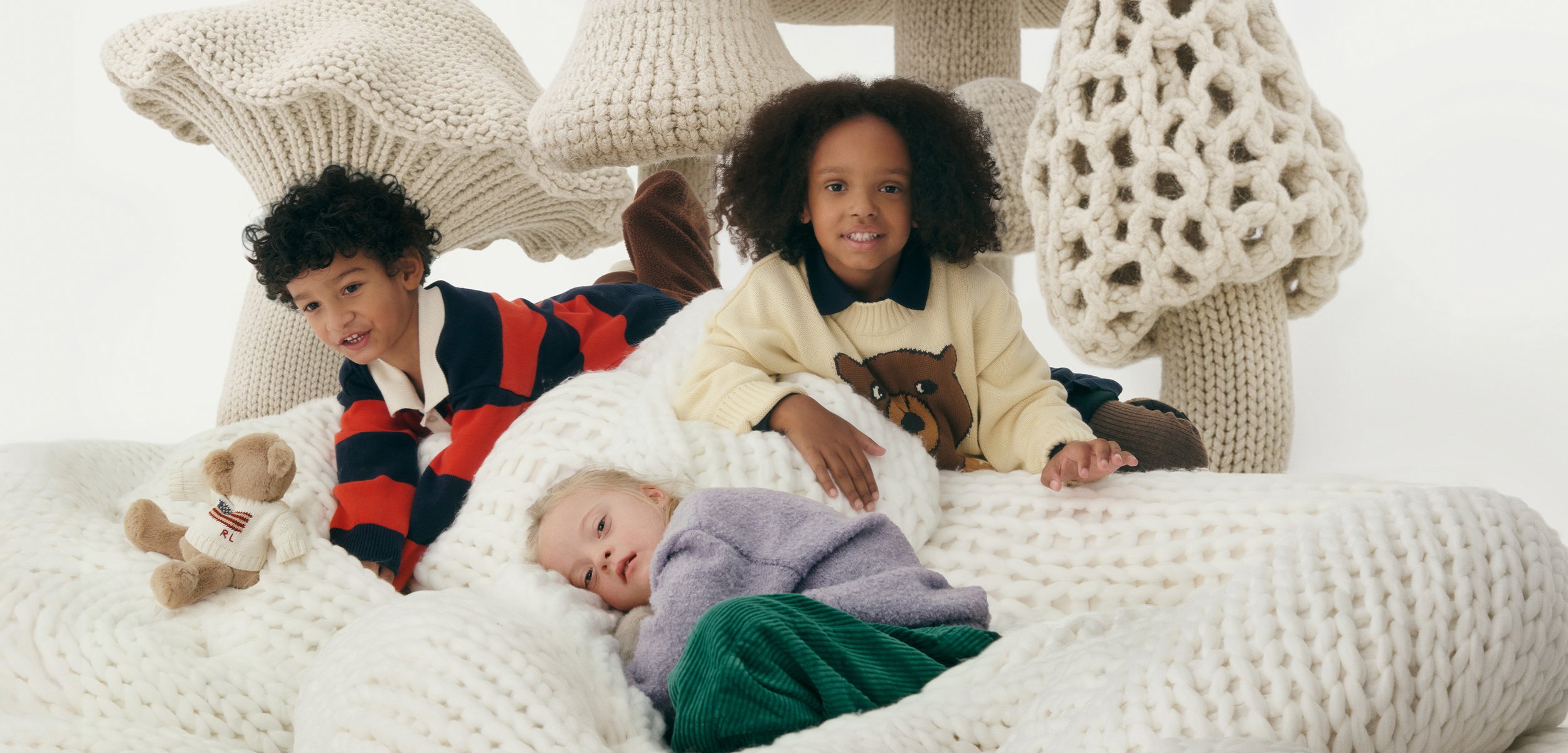 Tre børn ligger og sidder på et stort hvidt chunky strikket tæppe med overdimensionerede strikkede svampe-skulpturer bag dem.
