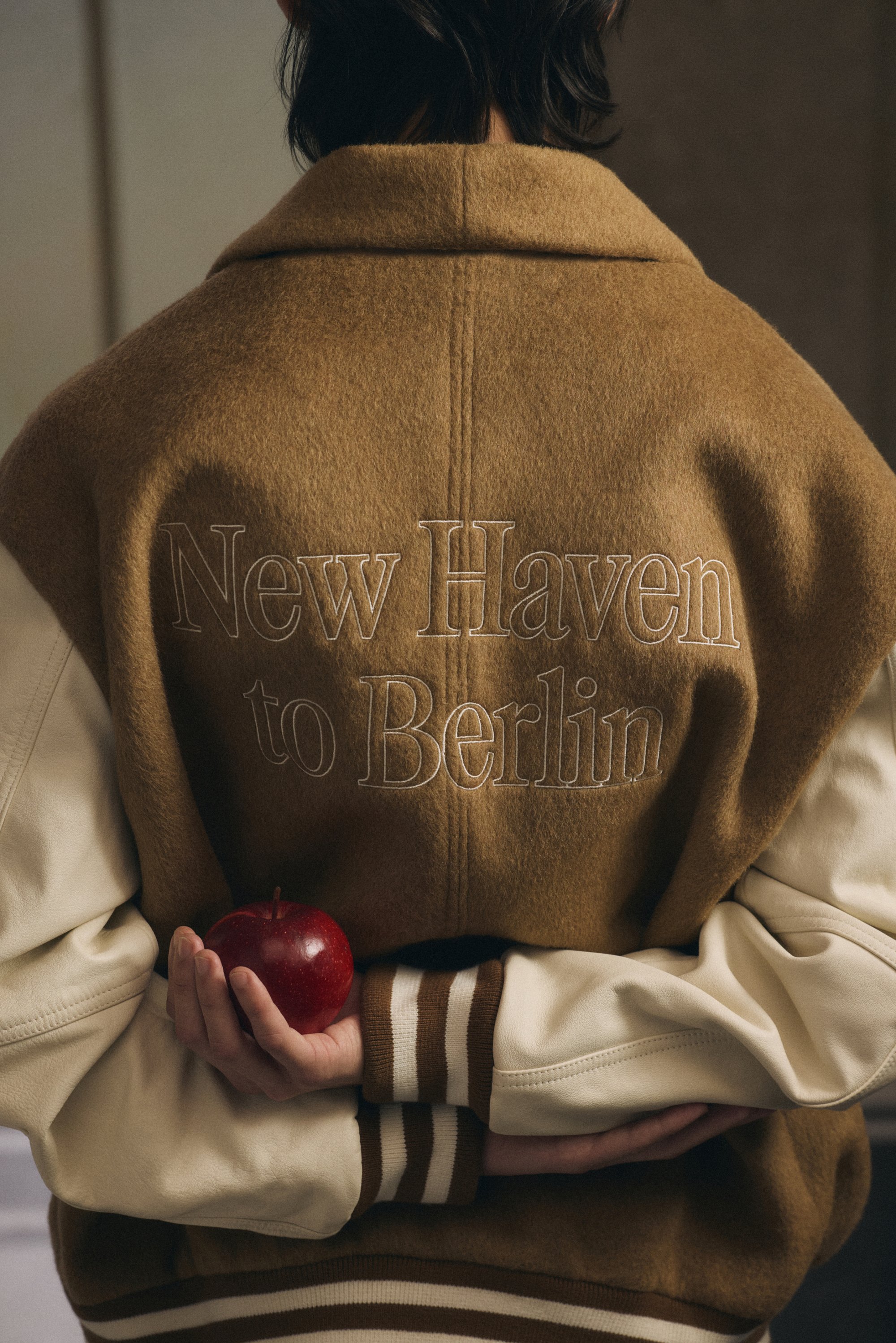 En person håller en röd äpple med båda händerna, står med ryggen mot kameran, iklädd en brun och beige jacka med "New Haven to Berlin" broderat.