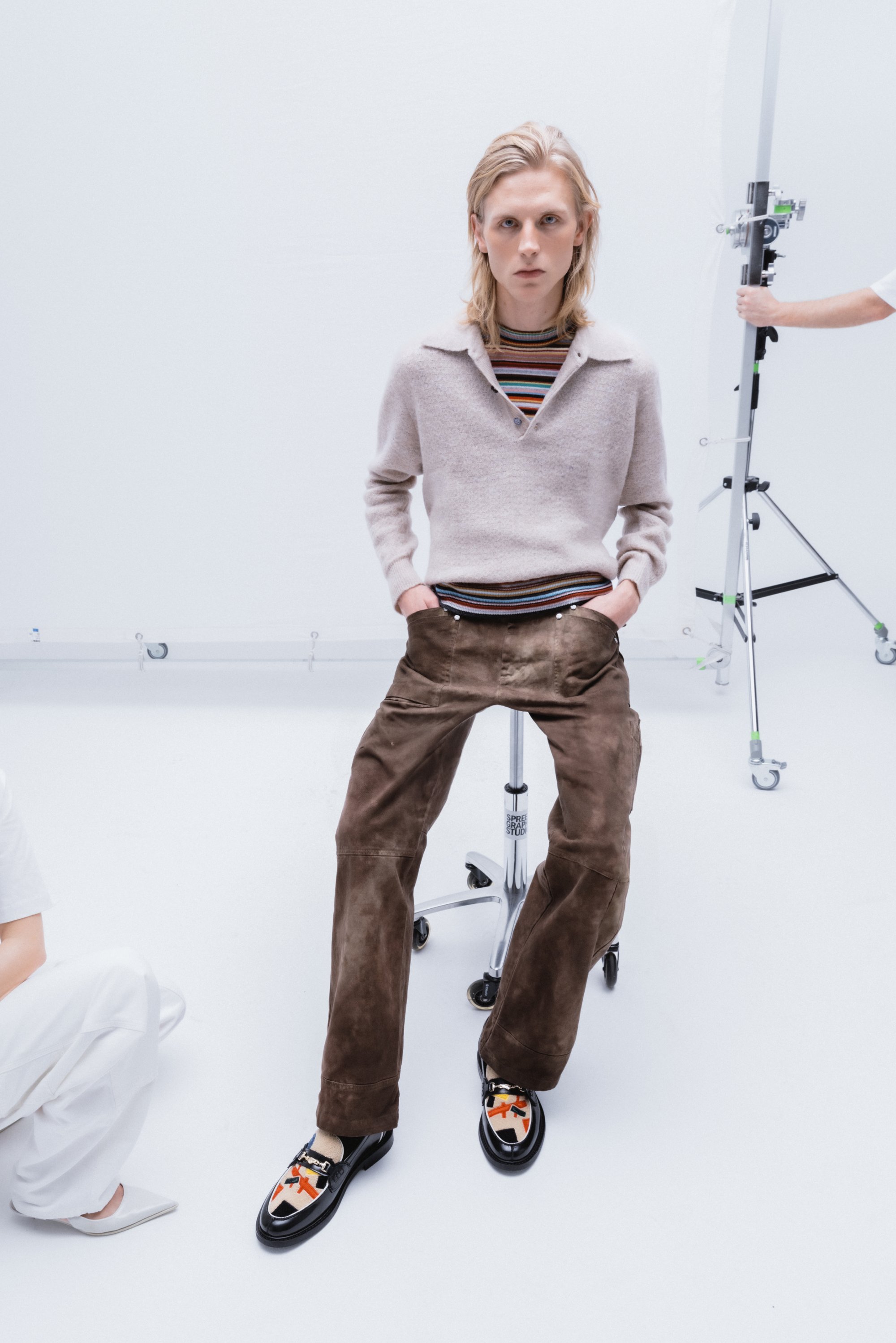 Una joven modelo con cabello rubio largo lleva un suéter claro y pantalones marrones, sentada en un taburete con ruedas en un luminoso estudio.