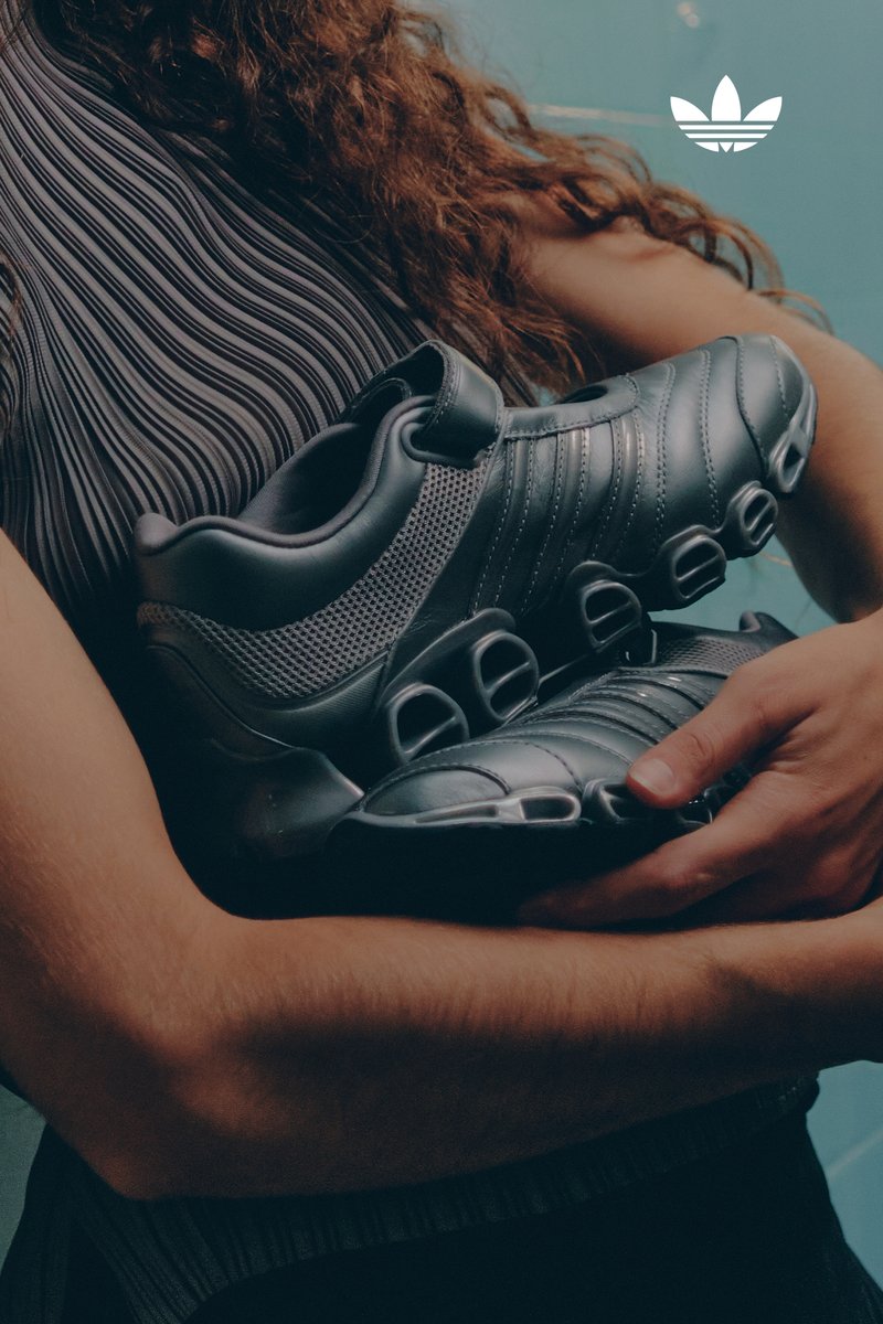 A person with curly hair cradles a pair of sleek, dark athletic shoes against a blue background. Adidas logo is visible.