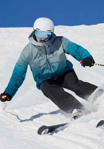 Ο εξοπλισμός σκι π�εριλαμβάνει ένα μπουφάν σε δύο χρώματα, τιρκουάζ και γκρι, μαύρα παντελόνια και γυαλιά σκι. Ο σκιέρ στρέφεται στο χιόνι με τα σκι του μερικώς βυθισμένα.