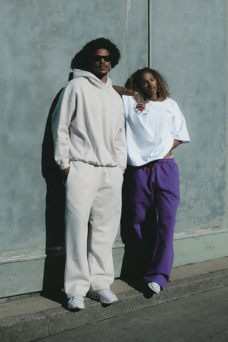 Due individui si appoggiano a un muro. Uno indossa un hoodie color crema e pantaloni abbinati; l'altro indossa una maglietta bianca e pantaloni viola.
