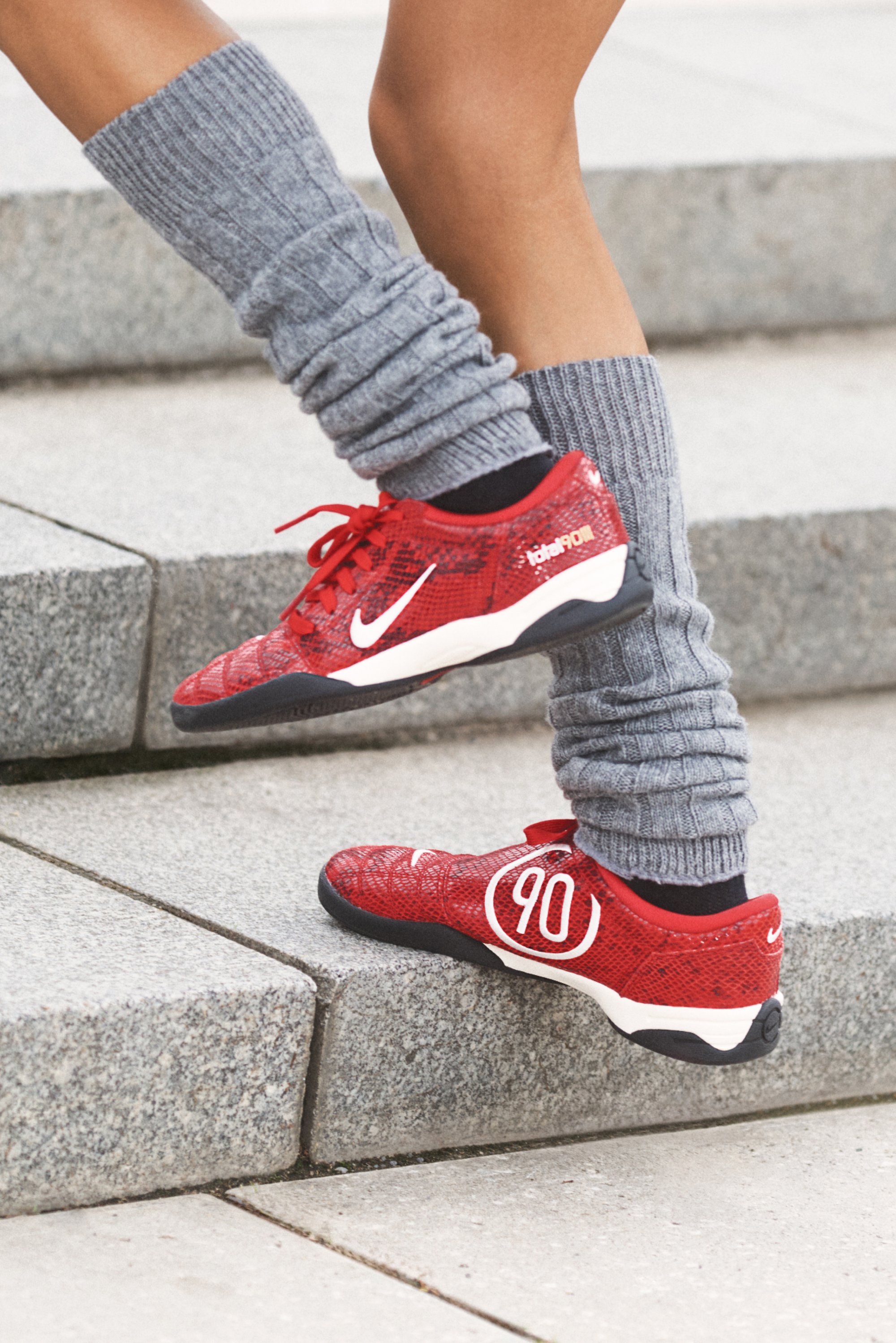 Zapatillas deportivas rojas con una superficie texturizada, que presentan un swoosh blanco y el número "90" en blanco, combinadas con calentadores de piernas grises con canalé.