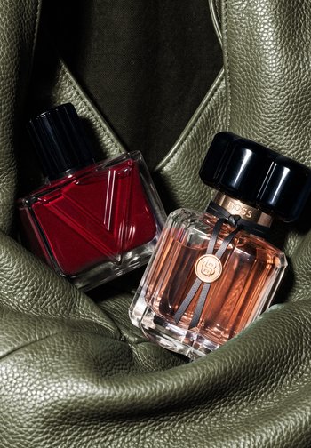 Deux flacons de parfum, un rouge avec un bouchon noir, un transparent avec un liquide pêche et un bouchon noir, reposant sur une surface en cuir vert olive texturée.
