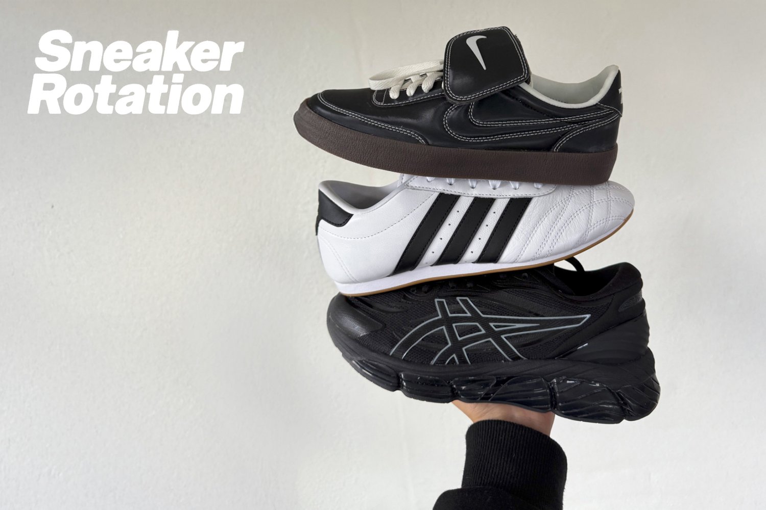 Een hand houdt een stapel van drie sneakers vast: zwarte Nike, witte Adidas en zwarte Asics, tegen een effen muur. "Sneaker Rotatie" is te zien.