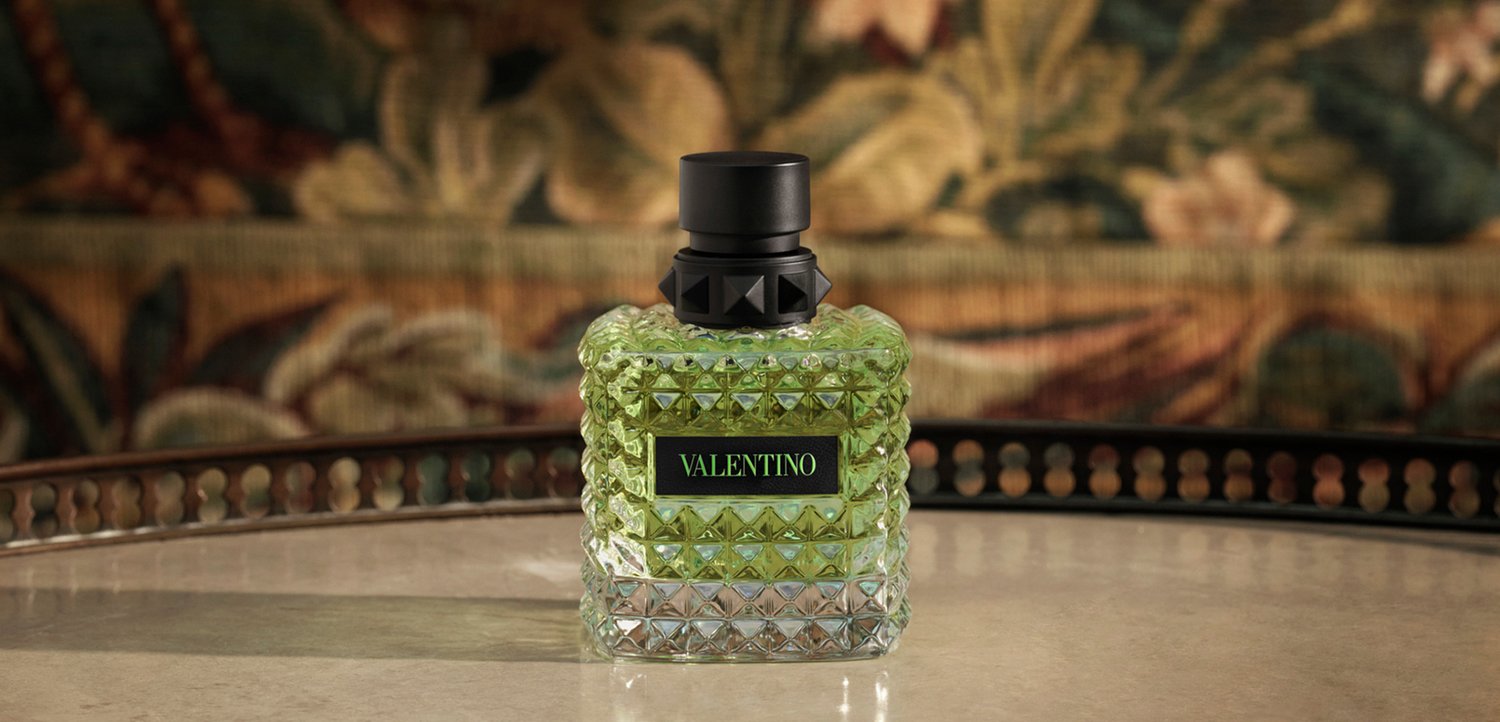 Vierkant getextureerde glazen parfumfles met Valentino-label en zwarte dop op reflecterend oppervlak en onscherpe bloemenachtergrond.