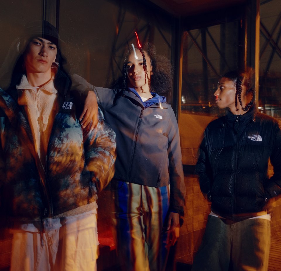 Three young individuals pose outdoors at night, wearing colorful North Face jackets, with a warm, urban backdrop and a soft, blurred effect.