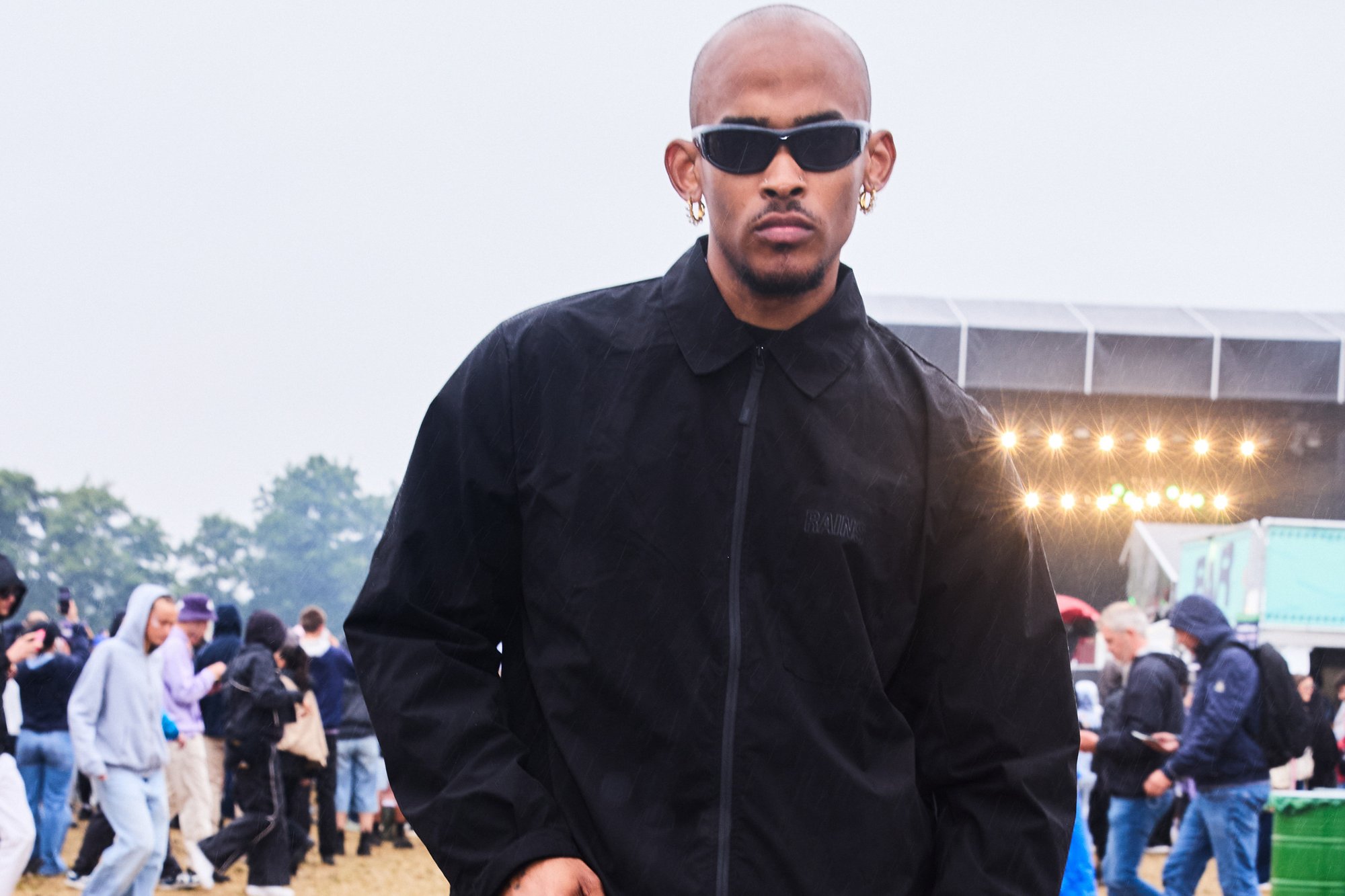 Veste coupe-vent noire avec un col montant, un ourlet incurvé et une fermeture éclair à l'avant. Portée avec des lunettes de soleil élégantes et des boucles d'oreilles en or à créole dans un cadre de festival.