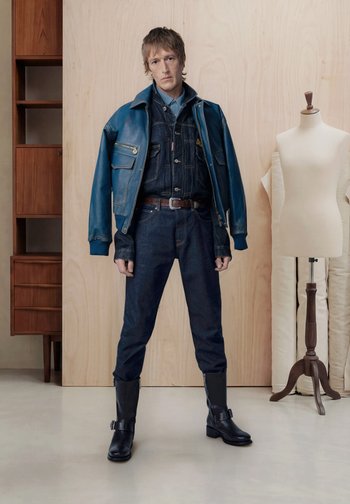 Man draagt gelaagde blauwe leren en denim jassen, denim broeken en zwarte laarzen staat binnen naast een houten kast en een paspop.