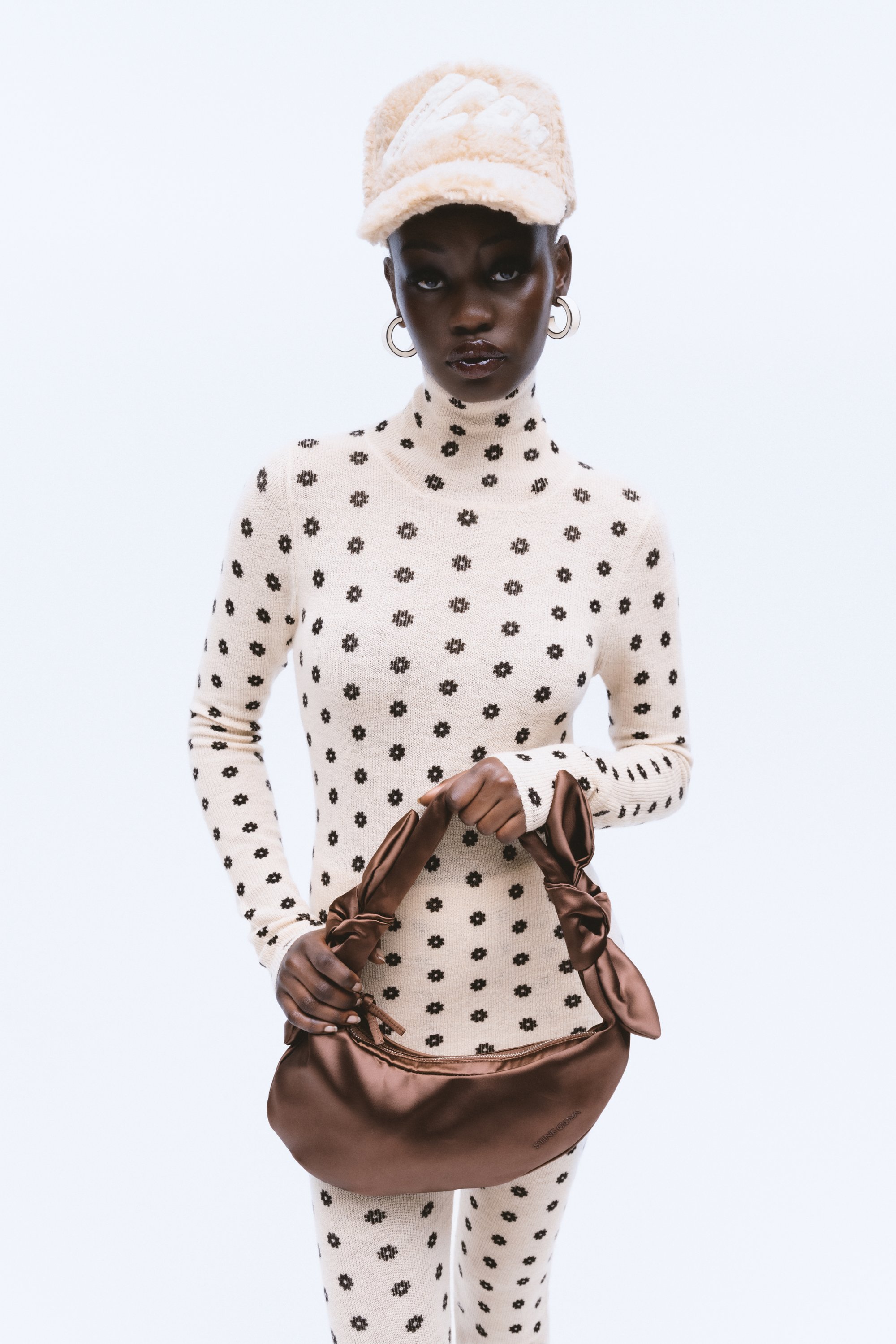 Model in a fitted cream outfit with black floral patterns, holding a brown satin handbag, wearing a fuzzy beige cap and hoop earrings.