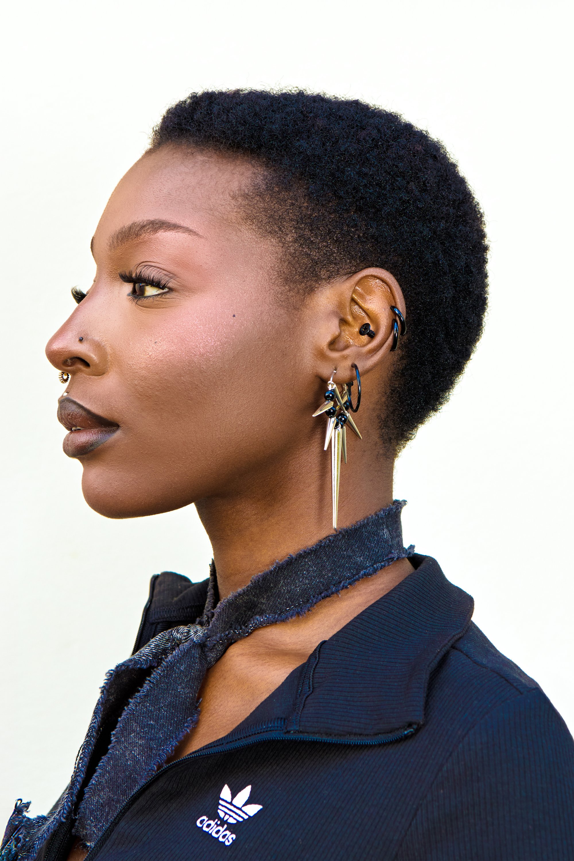 Guld spike øreringe med sorte accenter, båret med en sort jakke med en flosset krave. Kort, tekstureret mørkt hår fuldender looket.