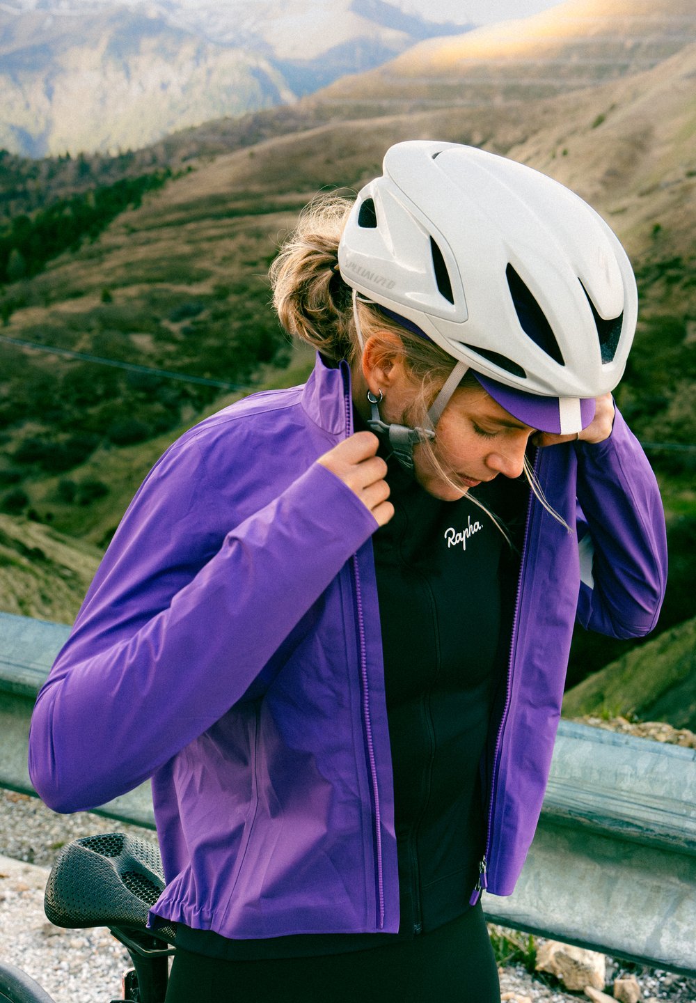 Sieviete, kas valkā baltu velosipēda ķiveri un purpura krāsas jaku, aizdara jakas rāvējslēdzēju, kamēr fona kalnu ainava.