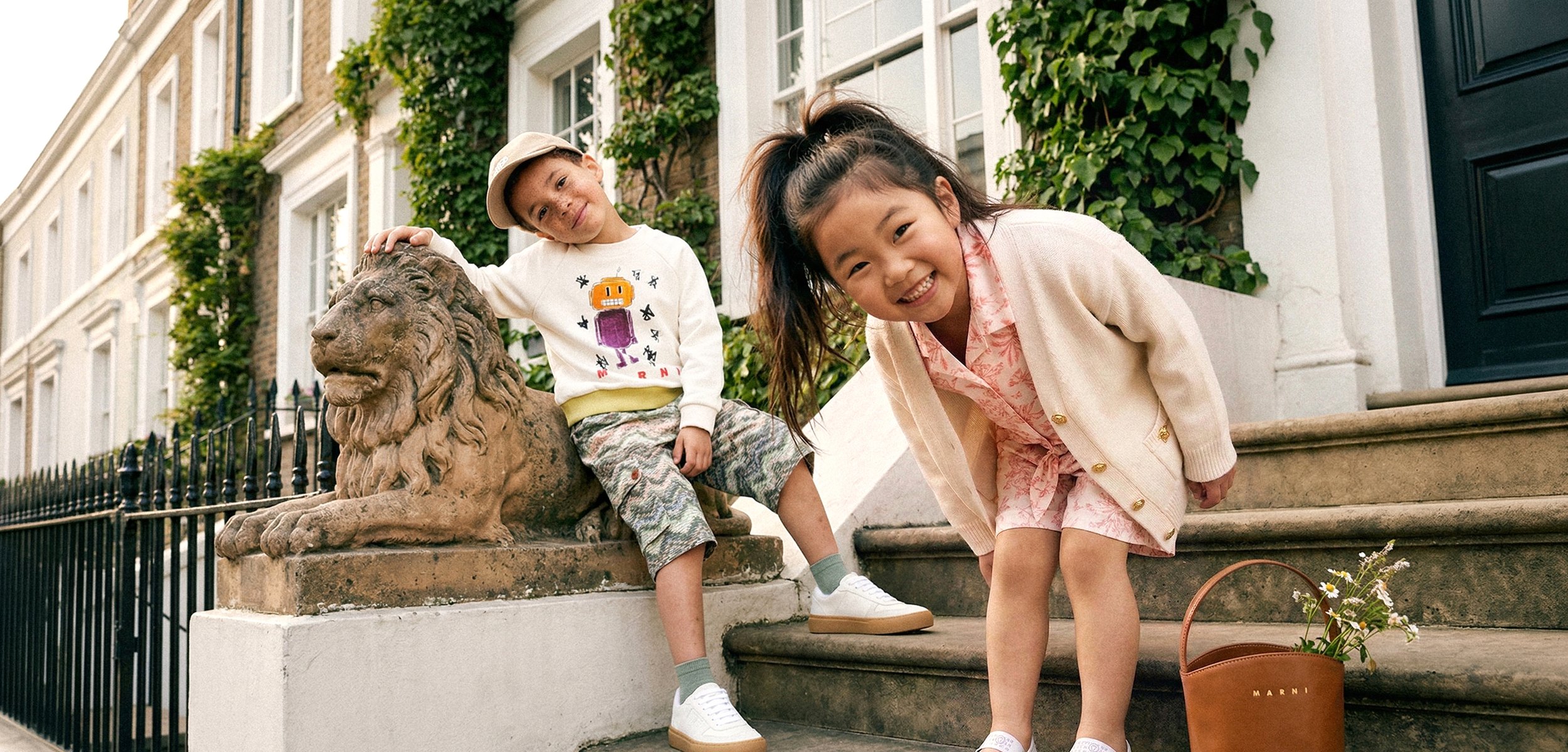 Zwei Kinder lächeln draussen; ein Junge lehnt an einer Löwenstatue, ein Mädchen beugt sich bei Steinstufen mit einem Korb Blumen vor.