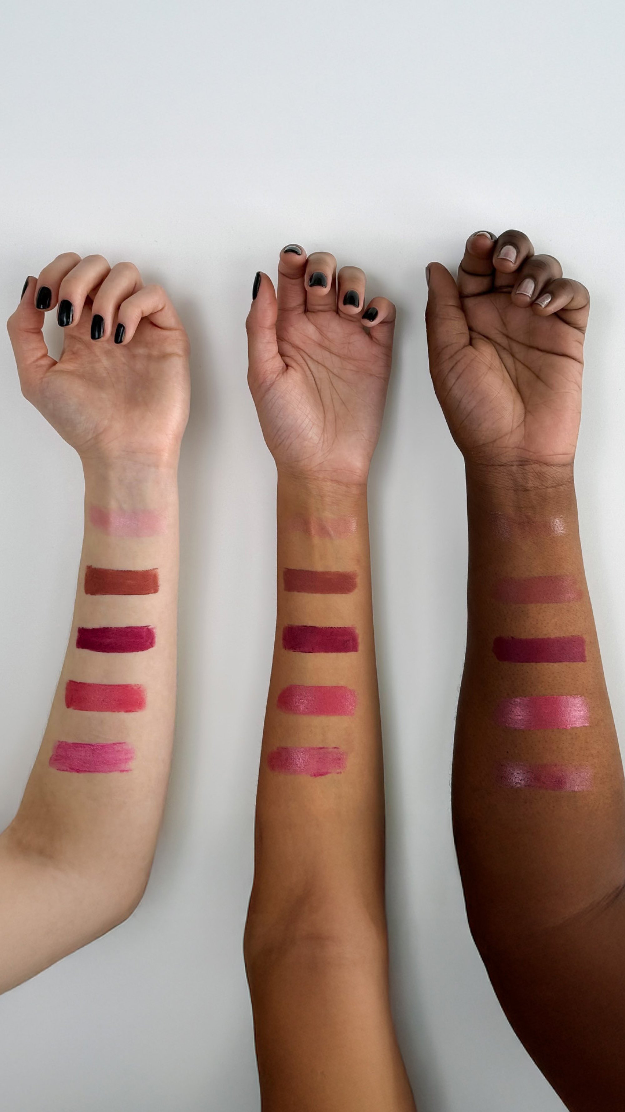 Tre braccia con campioni di rossetto: tonalità di pelle chiara, media e scura. I campioni includono rosa, rossi e mauve in varie finiture e texture.