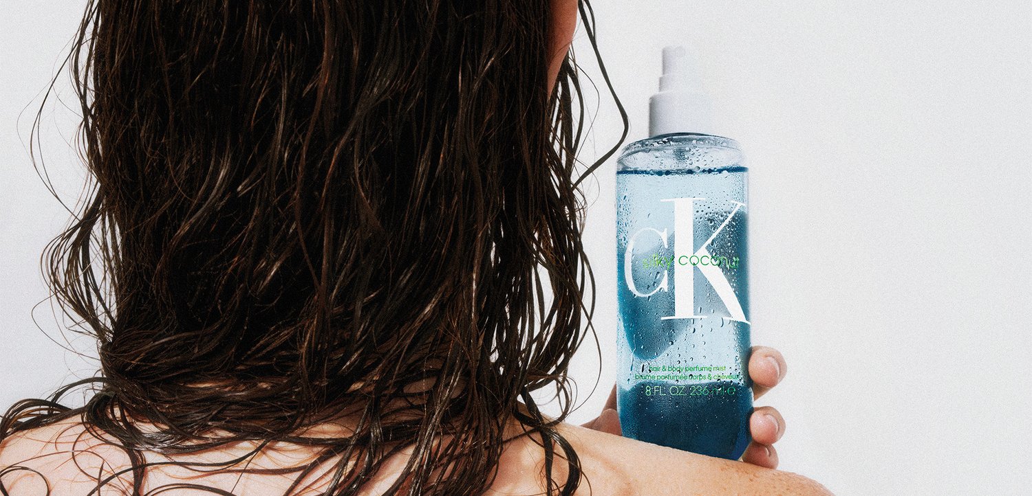 Person med vådt hår holder en blå CK Coconut shampoo flaske mod en hvid baggrund, der viser etiketten og væsken indeni.