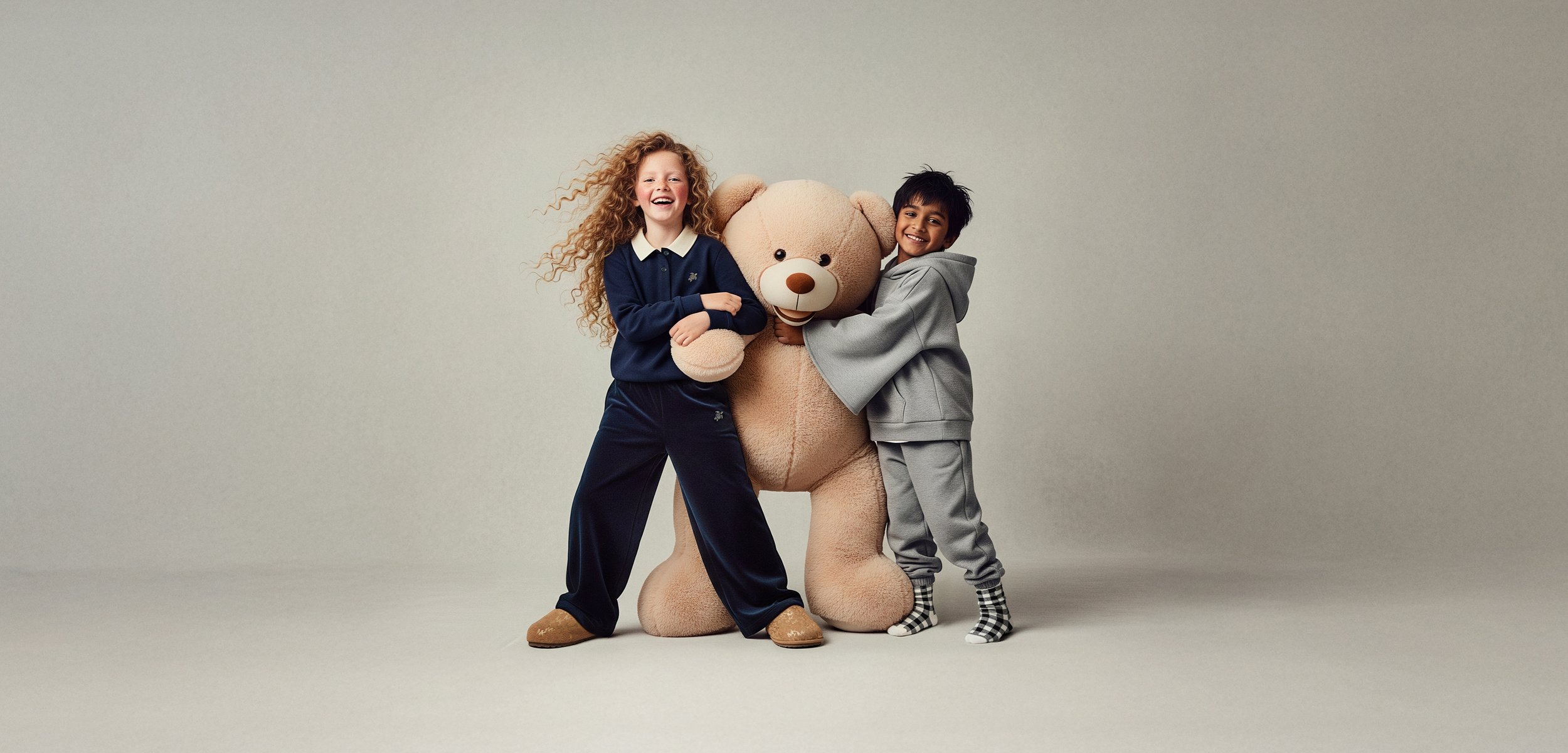 Δύο παιδιά αγκαλιάζουν μια μεγάλη, αδιάβροχη μπεζ αρκούδα. Το ένα παιδί φοράει ένα σκούρο ναυτικό σύνολο με λευκό γιακά, ενώ το άλλο είναι ντυμένο με γκρίζα ρούχα.