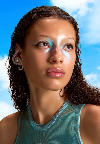 Une jeune femme aux cheveux bouclés et à la peau éclatante regarde vers le haut, portant un maquillage irisé sur les yeux et un haut bleu tendance sur un fond de ciel lumineux.