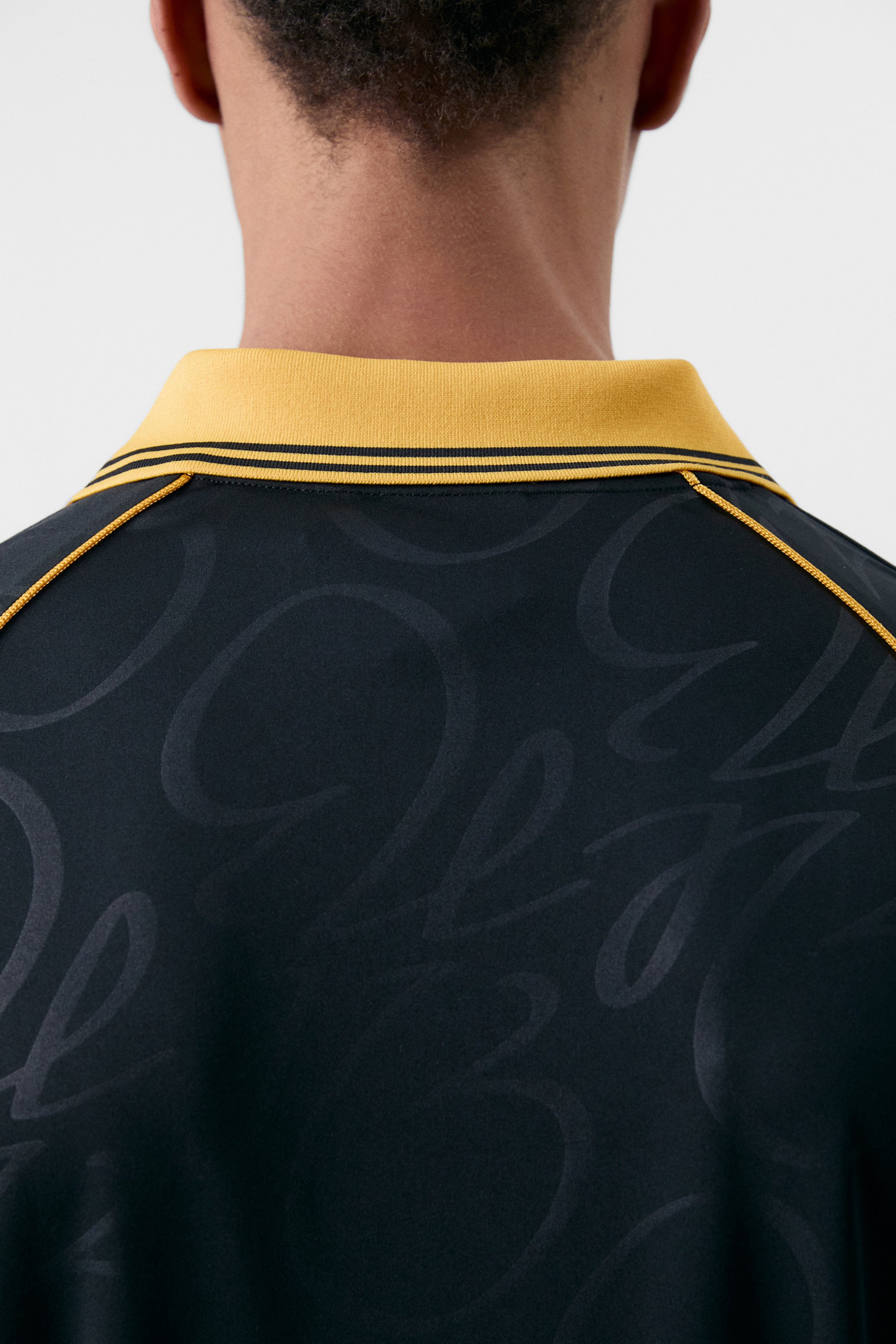 Acercamiento al cuello y espalda de una persona, que lleva una camisa polo negra con un collar amarillo y sutiles detalles en patrón.