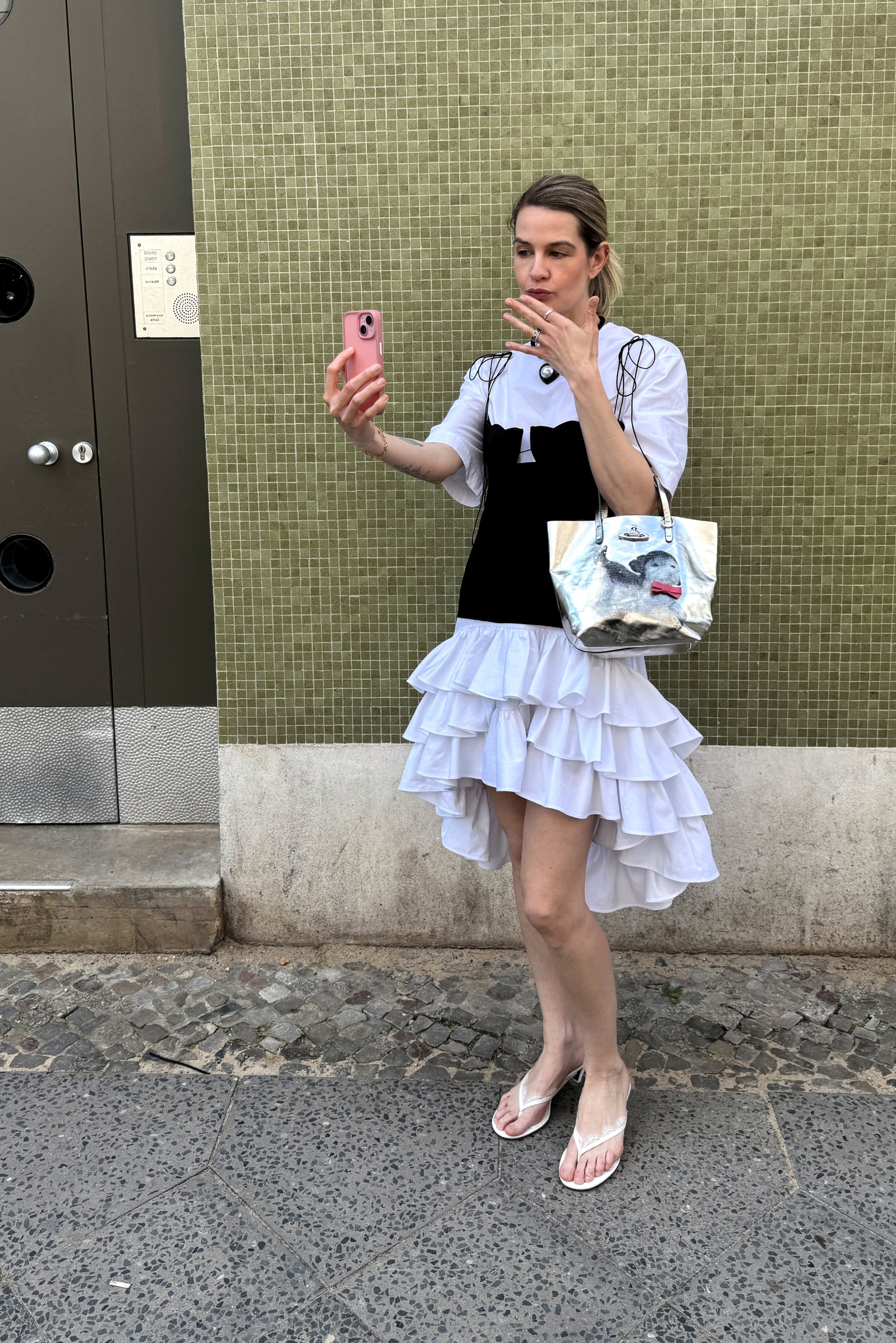 A person takes a selfie while wearing a black and white layered dress and holding a silver handbag, standing against a green tiled wall.