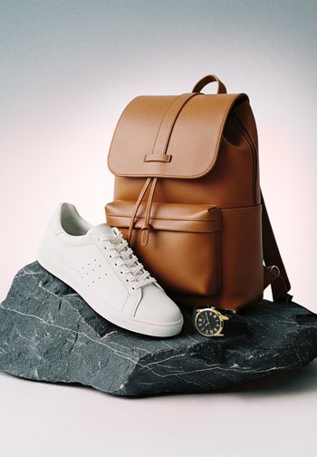 Pruun nahast seljakott, valged tossud ja must randmekell kuldsete detailidega tumedal kiviplaadil.