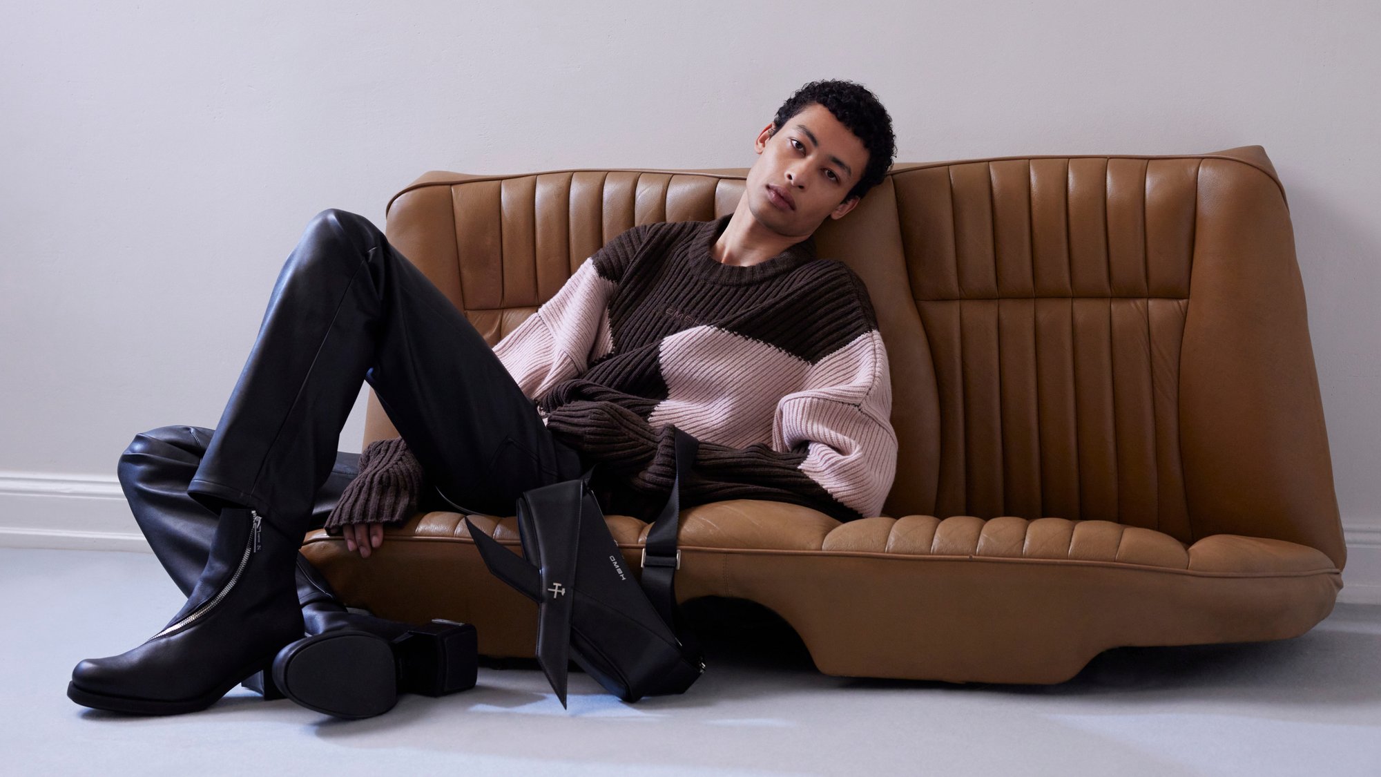 Una persona con un maglione a righe si rilassa su un divano retrò marrone, indossando pantaloni neri e stivaletti eleganti, con una borsa alla moda accanto a sé.