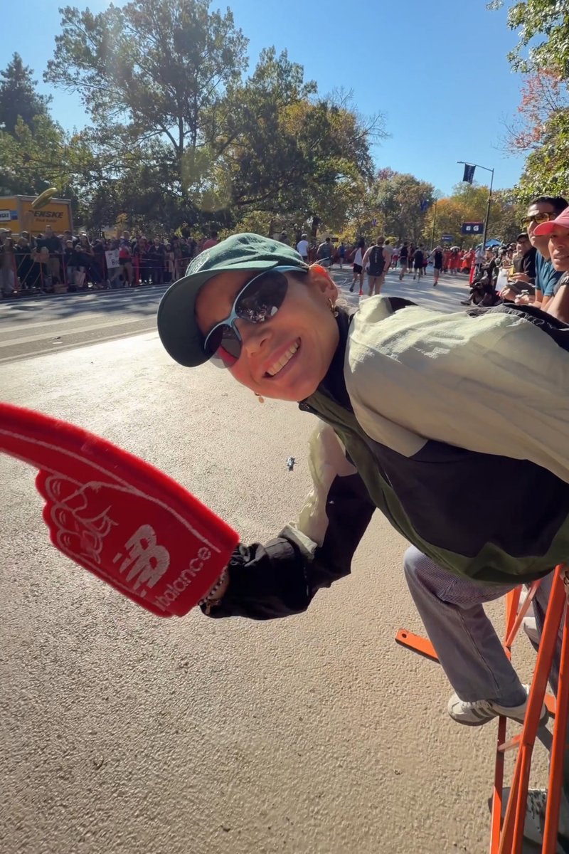 Lächende Person mit Sonnenbrille und Mütze hält einen roten Schaumstofffinger und feuert an, nahe einer Rennstrecke mit Läufern und einer Menschenmenge hinter Absperrungen.