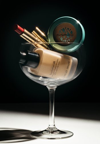 Clear glass bowl with a tall stem holds a foundation bottle, a green compact, and several lipsticks, including a red one.