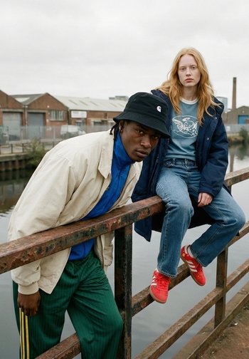 Dos jóvenes junto a la barandilla de un canal; uno se inclina hacia adelante con un sombrero negro y una chaqueta beige, el otro está sentado con una chaqueta azul y zapatillas rojas.