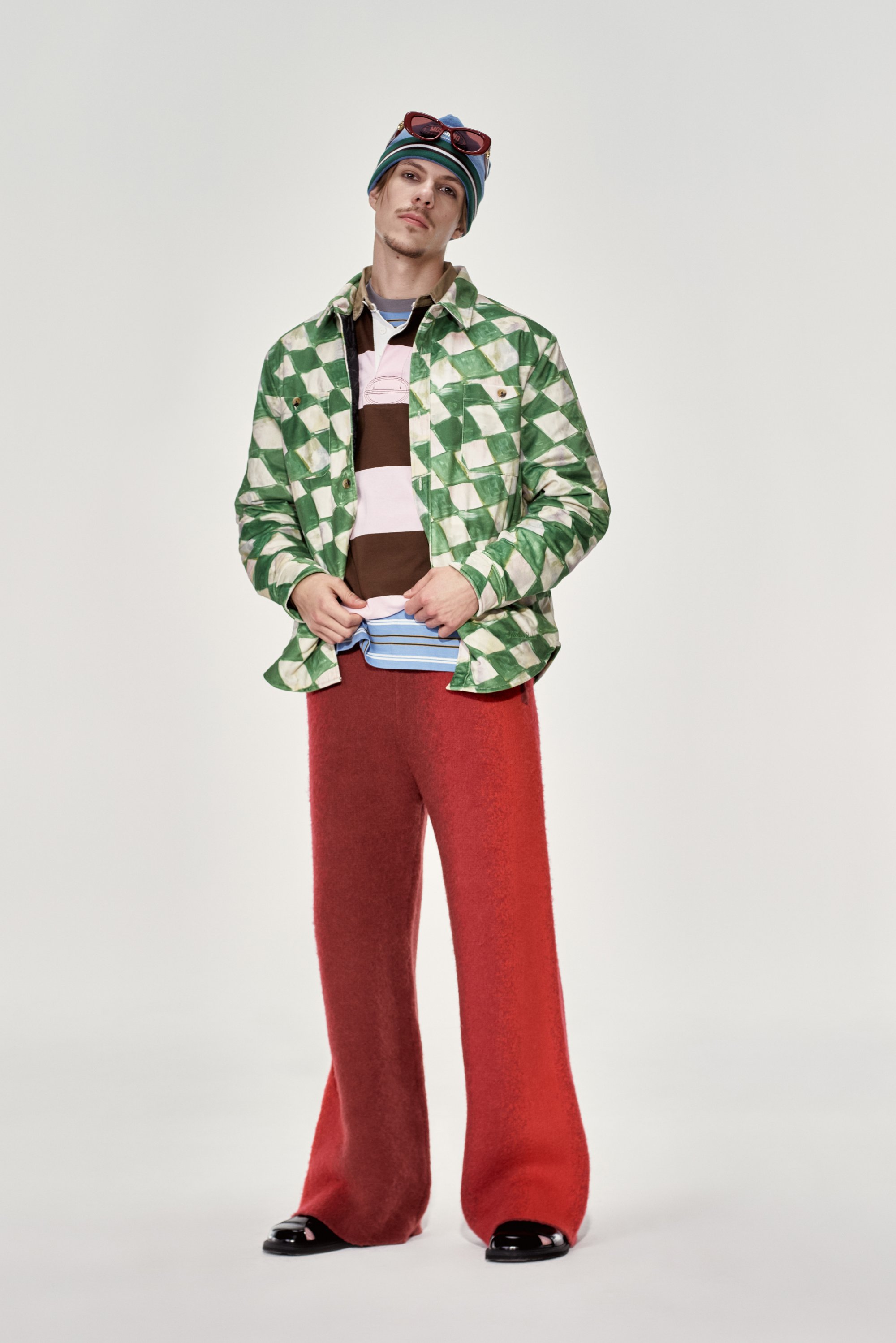Un hombre sostiene una expresión neutral, viste una chaqueta a cuadros verdes, camisas en capas y pantalones anchos de color rojo brillante. Lleva unas gafas de sol sobre su gorro.
