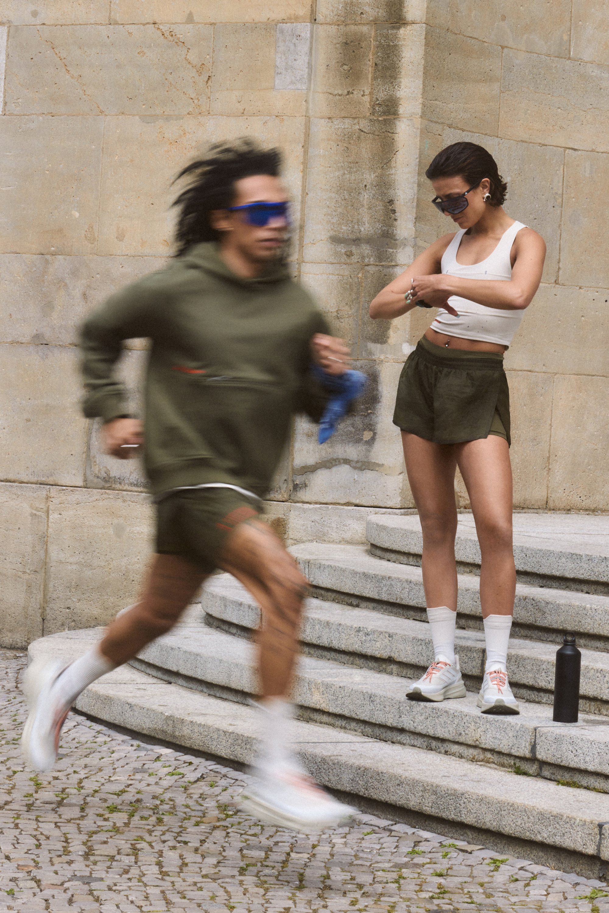 Un uomo scende correndo le scale di pietra indossando un hoodie verde e pantaloni corti, mentre una donna sta vicino a lui regolando il suo orologio, entrambi in abbigliamento sportivo.
