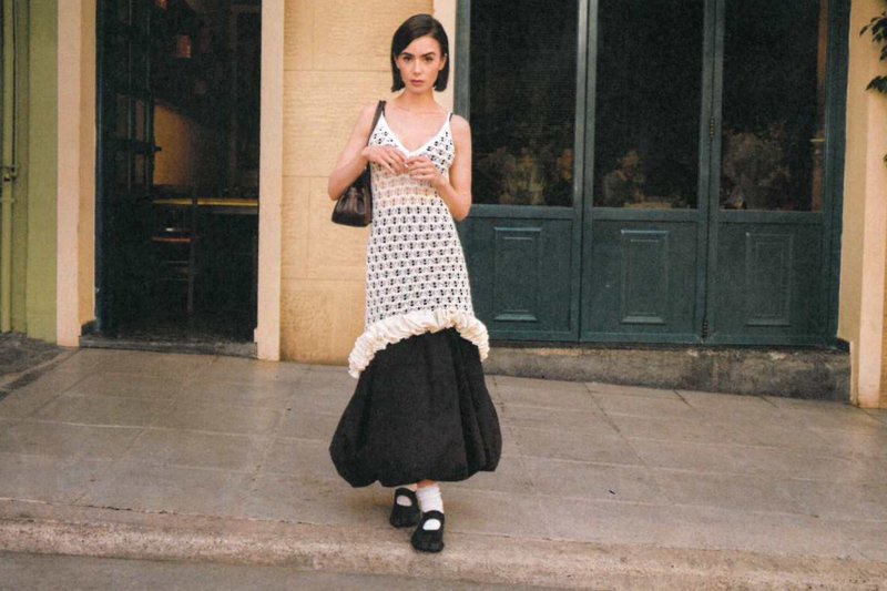 Junge Frau in einem schwarz-weißen Kleid mit Blumenmustern steht auf einem Bürgersteig vor grünen Doppeltüren.