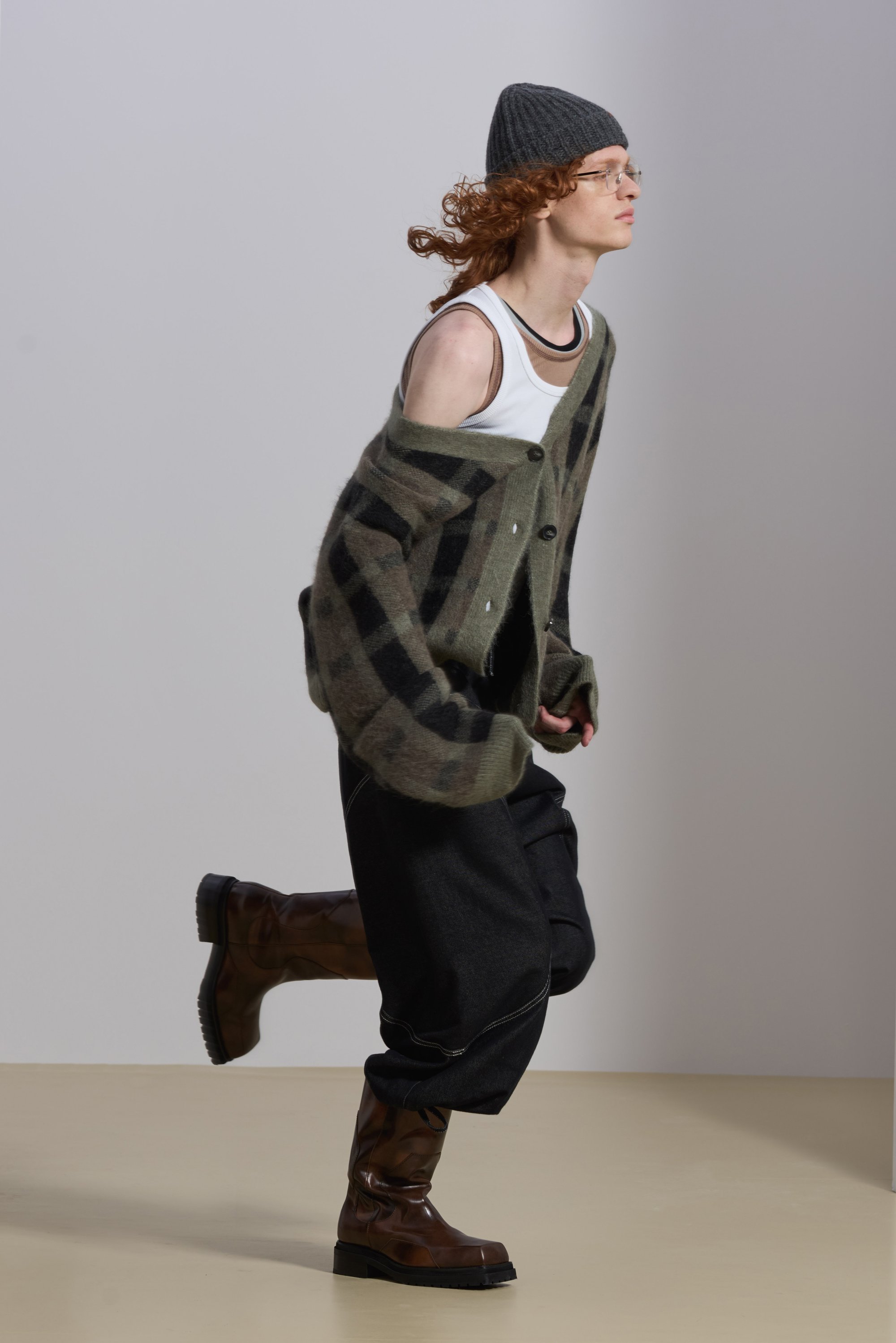 A person with curly red hair wears a plaid cardigan, tank top, and brown boots, mid-stride against a light background.